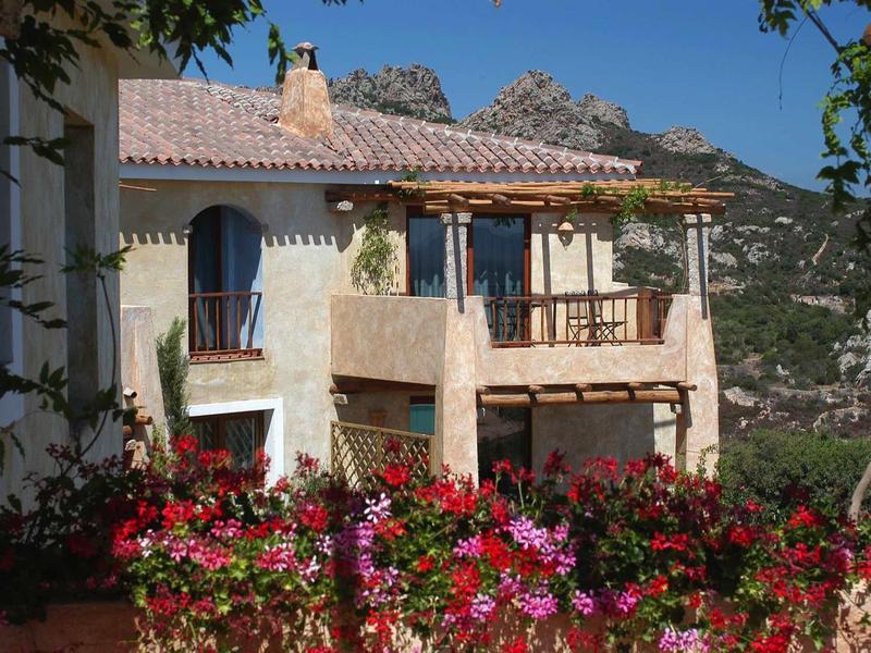 Weißes Haus mit rotem Blumengarten und Bergen im Hintergrund unter blauem Himmel.