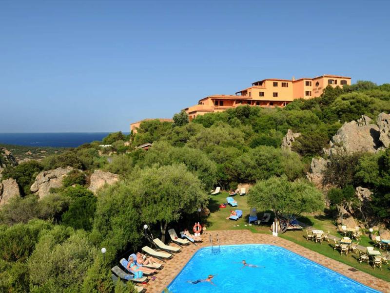 Hotel mit rötlichen Gebäuden auf grünem Hügel, blauem Himmel und rundem Pool im Vordergrund.