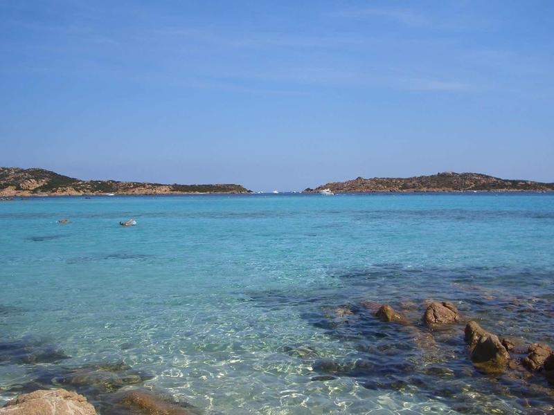 Klares blaues Meer trifft auf felsige Küste unter hellem Himmel mit vereinzelten Wolken.