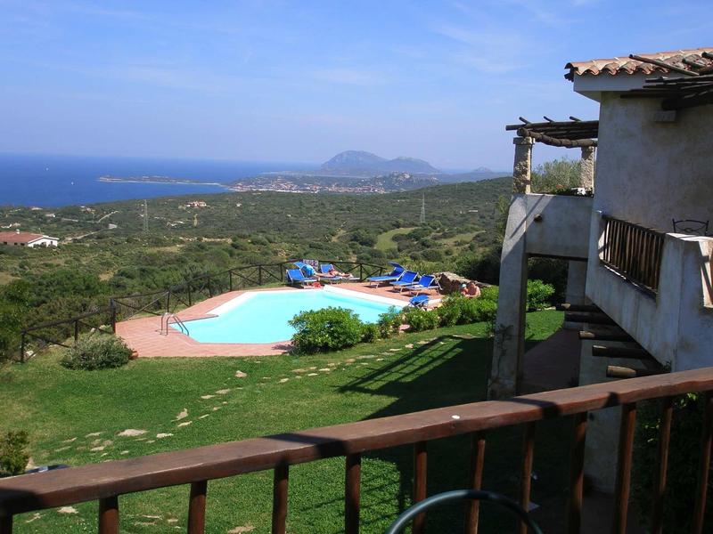 Blick von Balkon auf grüne Landschaft mit Pool, blauen Liegen und distantem Meer unter blauem Himmel.