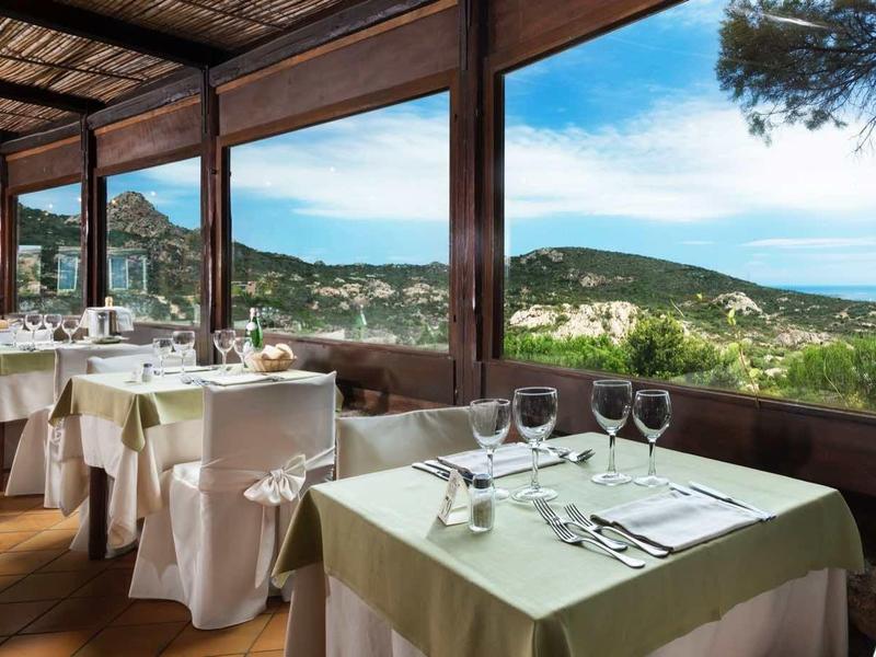 Restaurant mit gedeckten Tischen, Holzboden, großen Fenstern und Blick auf grüne Hügel und Meer.