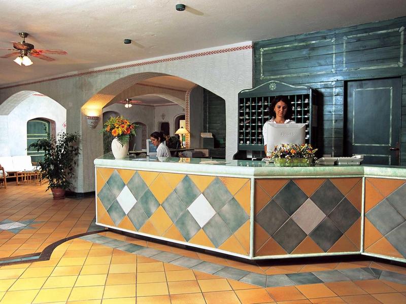 Hotelrezeption mit gemustertem Tresen, freundlicher Dame, warmen Fliesen und bogenförmigen Durchgängen.