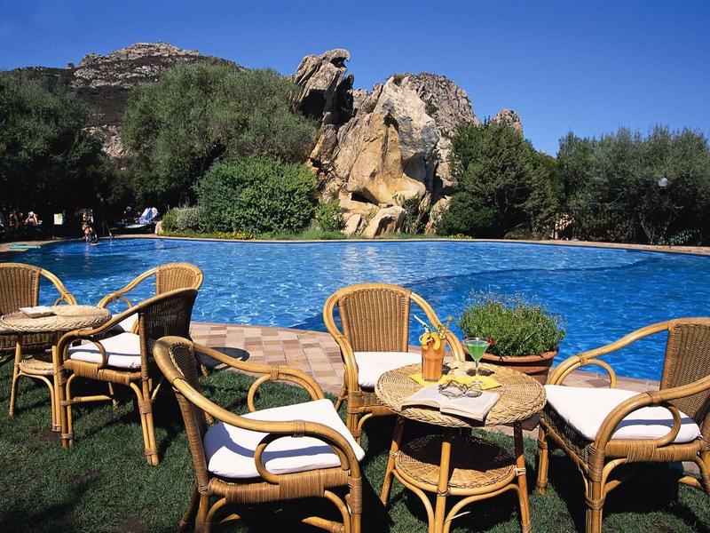 Runder Tisch mit vier Korbstühlen am Pool, dahinter Felsen, Bäume und klarer blauer Himmel.