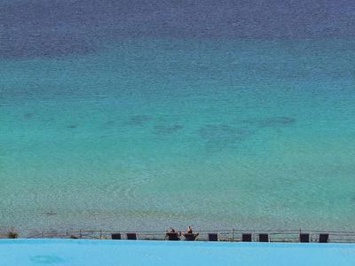 Ein großer Pool mit blauen Liegestühlen und Sonnenschirmen, dahinter ruhiges Meer und klarer Himmel.