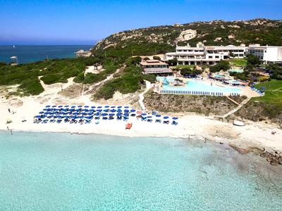 Strand mit klarem türkisfarbenem Wasser, blauen Sonnenschirmen und einem Hotel in Hügelnähe.