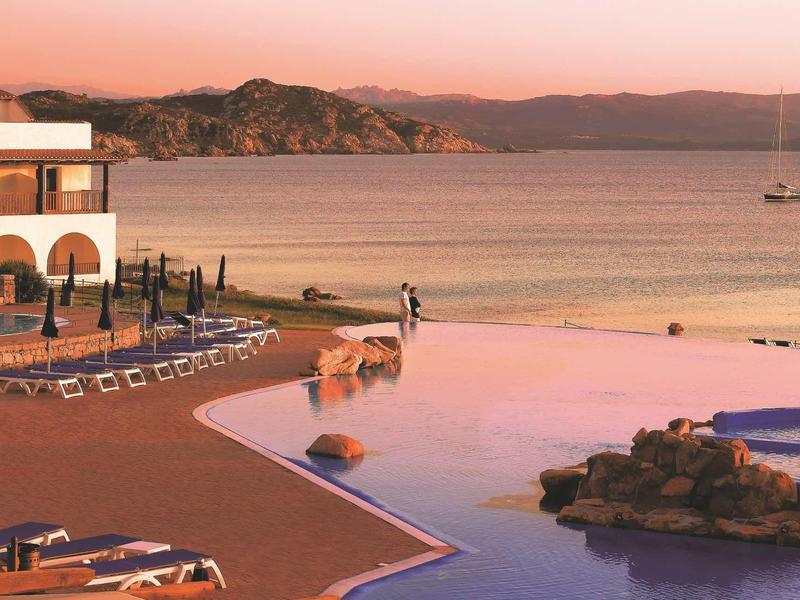 Infinity-Pool am Abend mit Blick auf ruhiges Meer und felsige Küste, Liegestühle und Gebäude links.