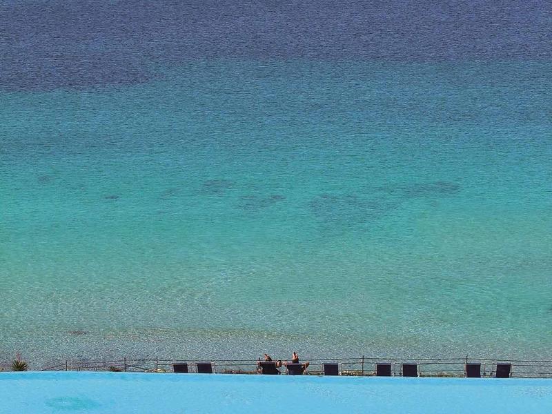 Ein großer Pool mit blauen Liegestühlen und Sonnenschirmen, dahinter ruhiges Meer und klarer Himmel.