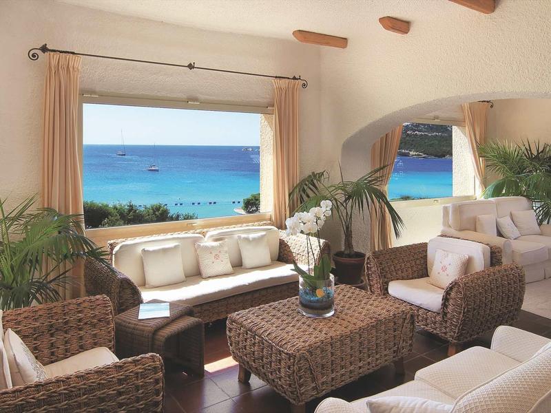 Helles Wohnzimmer mit Korbmöbeln, Pflanzen und Blick auf das blaue Meer durch ein großes Fenster.