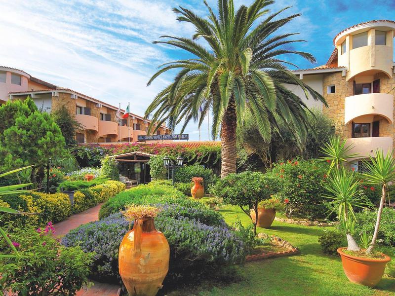 Hotel mit mediterranem Garten, Palmen und großen Tontöpfen vor dem Gebäude.