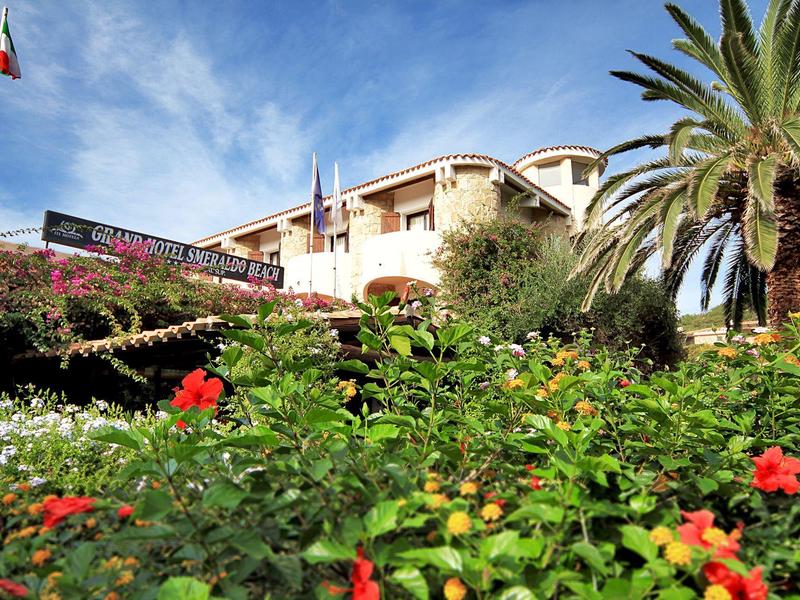Hotelgebäude mit Palmen und blühenden Pflanzen im Vordergrund bei blauem Himmel