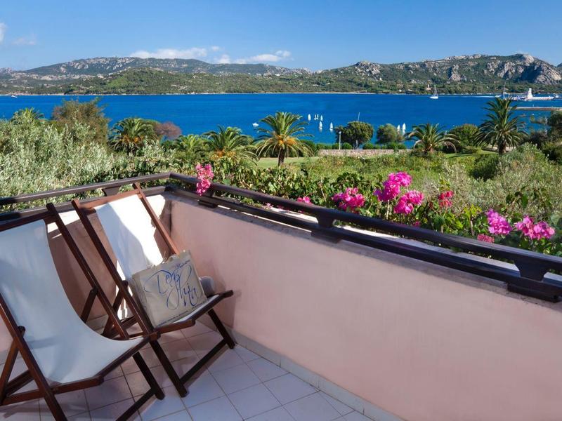 Balkon mit zwei Stühlen, pinken Blumen am Geländer, Blick auf blaues Meer und Hügel im Hintergrund.