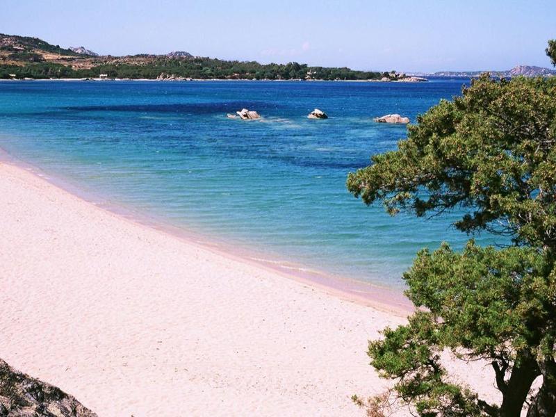Strand mit hellem Sand, blauem Wasser und grün bewachsenen Hügeln im Hintergrund.