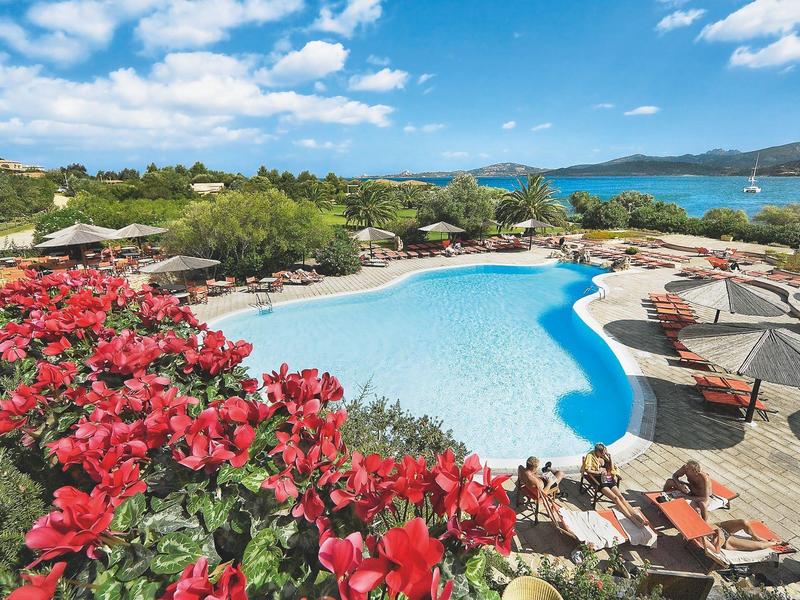Ein großer Pool mit blauen Wasser, umgeben von roten Blumen, Sonnenschirmen und grüner Natur am Meer.