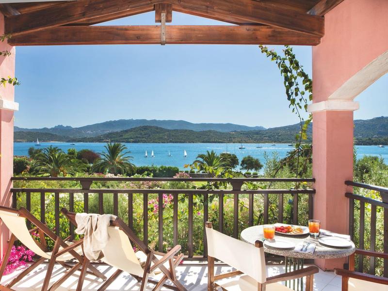 Balkon mit Holzmöbeln, pinken Wänden, Blick auf Palmen und azurblaues Meer unter blauem Himmel.