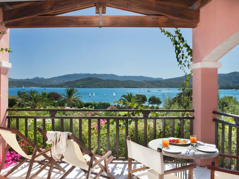 Balkon mit Holzgela?nder, Tischen, Stühlen und Meerblick mit grünem Ufer und Bergen im Hintergrund.