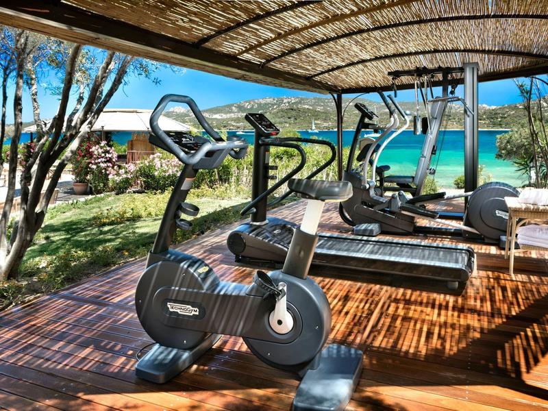 Freiluft-Fitnessbereich mit Fahrradtrainer und Laufbändern unter schattigem Holzdach, grüne Natur.