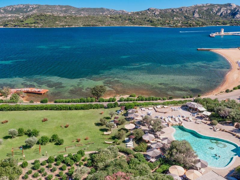 Blick auf türkisfarbenes Meer, Sandstrand, grünes Gelände und Swimmingpool mit Sonnenschirmen.
