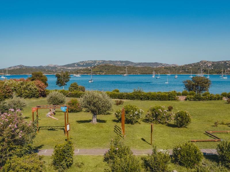 Grüne Wiese mit Büschen, dahinter blauer Fluss und hügelige Landschaft unter klarem Himmel.