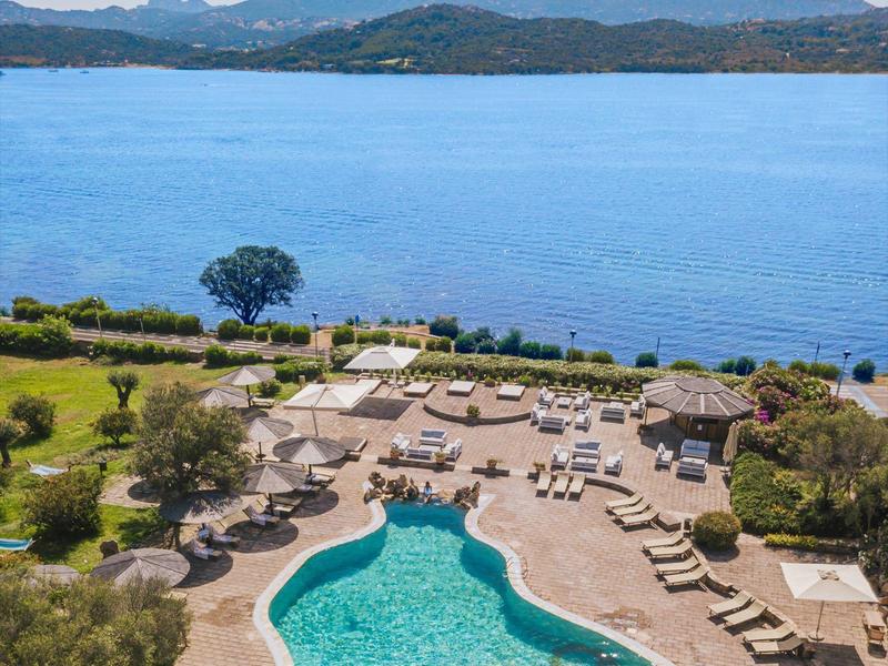 Pool mit unregelmäßiger Form, umgeben von Liegestühlen, Palmen und Blick auf blauen See und Berge.