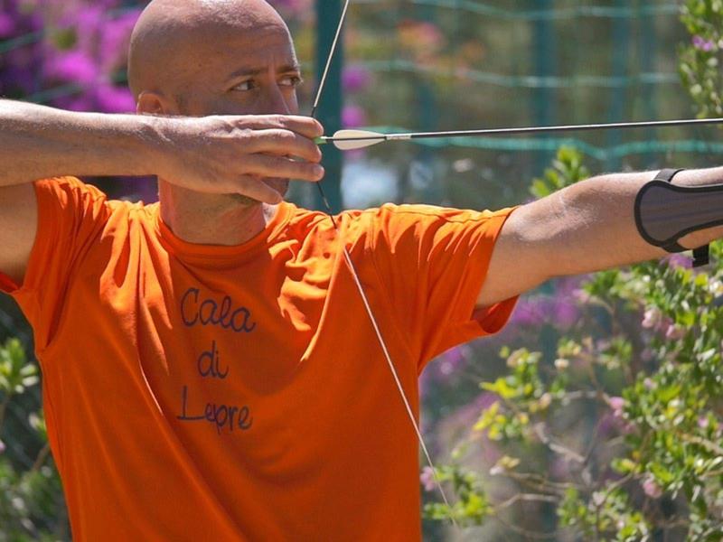 Mann in orangem T-Shirt zieht Bogen mit Pfeil, grüner Hintergrund und Naturumgebung.