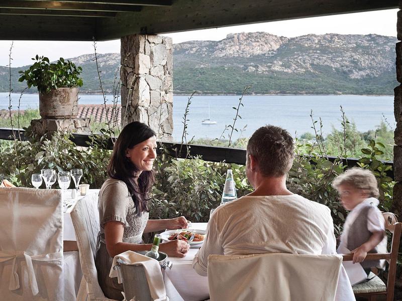 Drei Personen sitzen an gedecktem Tisch unter schattigem Dach mit Blick auf Wasser und Berge.