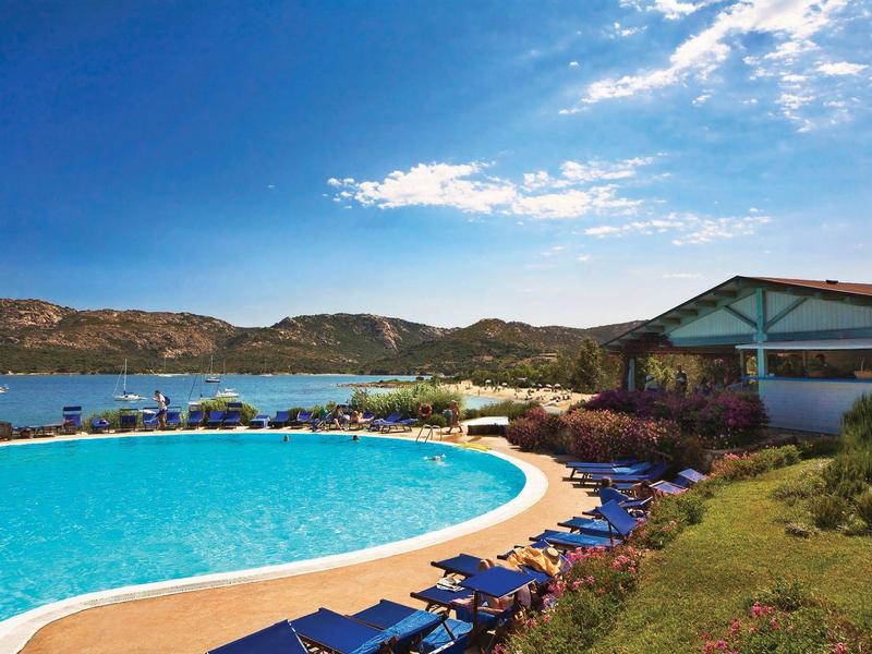Großer Außenpool mit blauem Wasser, Liegen, bewachsenem Hang und Blick auf Meer und Hügel.