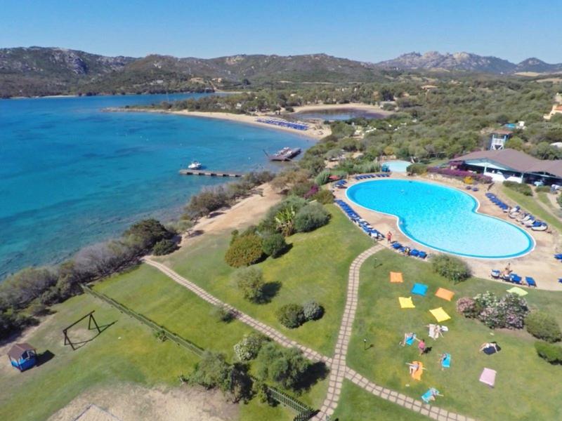 Küste mit blauem Meer, einem geschwungenen Schwimmbad und bunten Sonnenschirmen auf grünem Rasen.