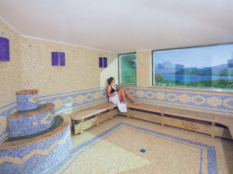 Raum mit Mosaikfliesen, Bank, und großer Fensterfront mit Bergblick, Frau sitzt entspannt darauf.