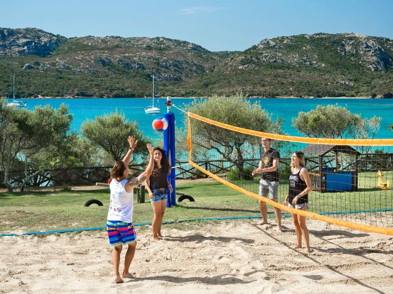 Vier Personen spielen Beachvolleyball auf einem Sandfeld mit Bergen und blauem Wasser im Hintergrund.