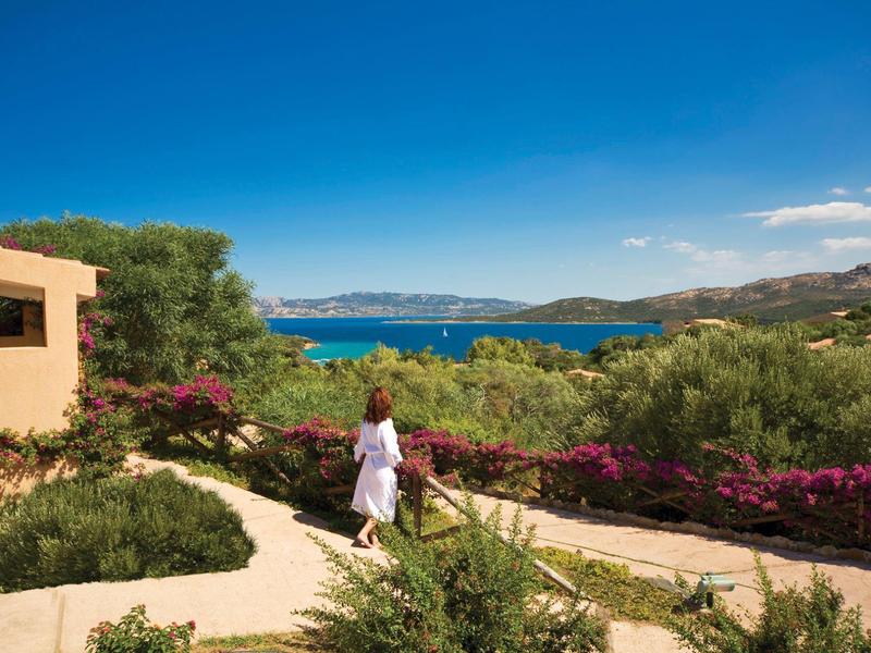 Frau in weißem Kleid wandert auf Plattenweg, umgeben von grünen Büschen und blühenden Pflanzen am Meer.