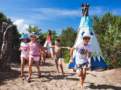 Kinder spielen draußen auf Sand vor bunten Indianerzelten unter blauem Himmel.