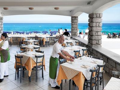 Strandrestaurant mit gedeckten Tischen, Kellnern in weißen Hemden und Meerblick im Hintergrund.