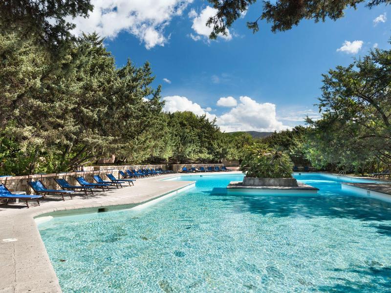 Großer blauer Swimmingpool mit blauen Liegestühlen und grünen Bäumen unter blauem Himmel.