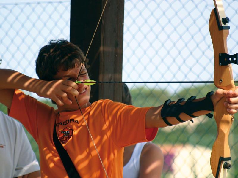 Junger Mann zielt mit Holzbogen, trägt orange Shirt und Armschutz, Hintergrund Drahtzaun und Himmel.