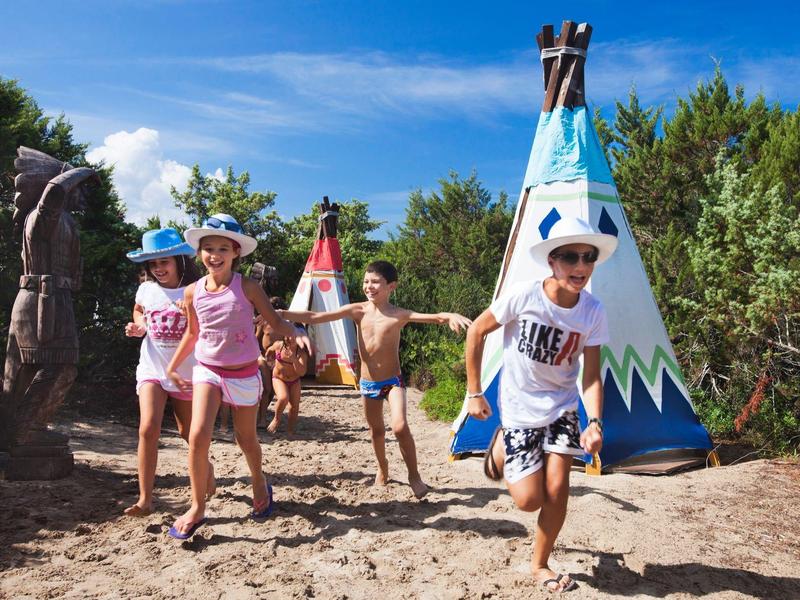 Kinder spielen draußen auf Sand vor bunten Indianerzelten unter blauem Himmel.