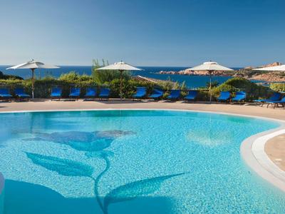 Rundlicher Pool mit blauem Wasser, blauen Liegen und weißen Sonnenschirmen am Meerufer.