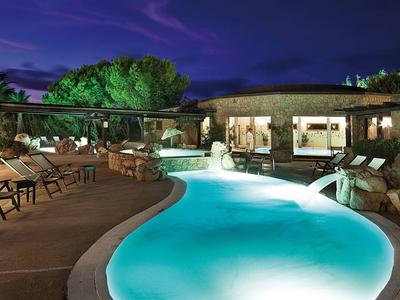 Beleuchteter Pool mit Wasserspielen, Außensitzbereich und Pflanzen bei Nacht.