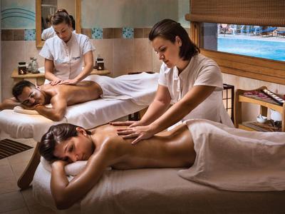 Zwei Frauen geben Rückenmassagen in einem warm beleuchteten Spa-Raum mit Holz und Meeresblick.