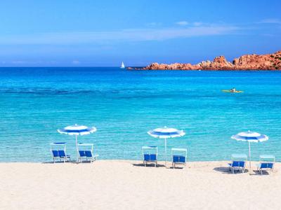 Weißer Sandstrand mit drei blauen Liegestühlen und Sonnenschirmen, türkisblaues Meer, heller Himmel.