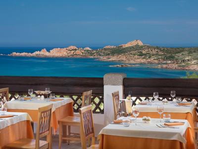 Restaurantterrasse mit gedeckten Tischen, Blick auf blaues Meer und felsige Küste bei klarem Himmel.