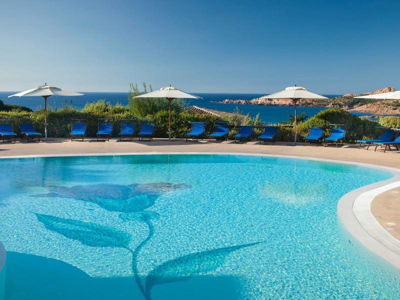 Ein großer, klarer Pool mit Blumenmuster, blauen Liegestühlen und Sonnenschirmen am Meer.