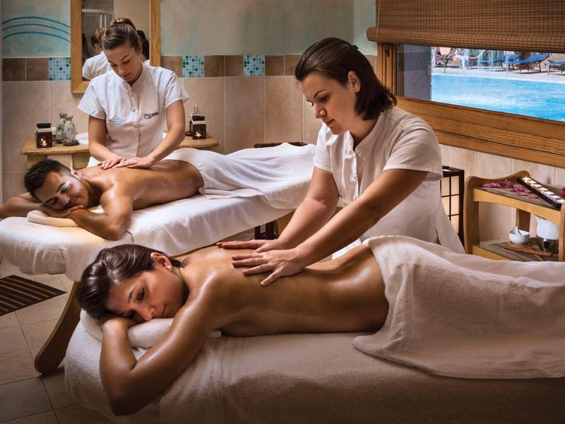 Zwei Frauen geben Rückenmassagen in einem warm beleuchteten Spa-Raum mit Holz und Meeresblick.