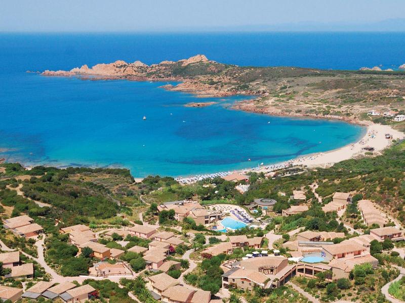 Küstenlandschaft mit türkisblauem Meer, Sandstrand und grüner Vegetation rund um Häuser.