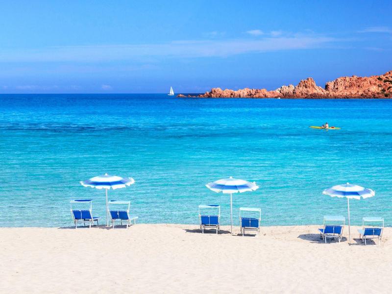 Weißer Sandstrand mit drei blauen Liegestühlen und Sonnenschirmen, türkisblaues Meer, heller Himmel.