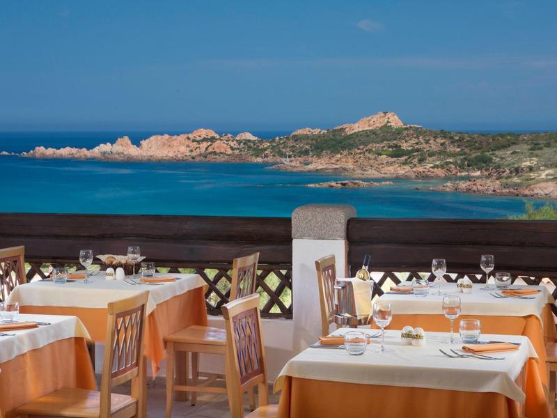 Restaurantterrasse mit gedeckten Tischen, Blick auf blaues Meer und felsige Küste bei klarem Himmel.