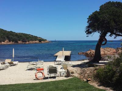 Strand mit Liegestühlen und klarem blauem Meer, umgeben von grünen Hügeln und Bäumen.