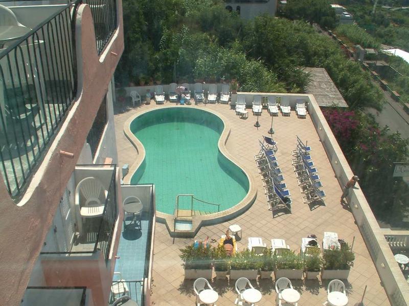Blick von oben auf einen Hotelpool mit Liegen und Tischen, umgeben von grünen Hügeln und Meer im Hintergrund.