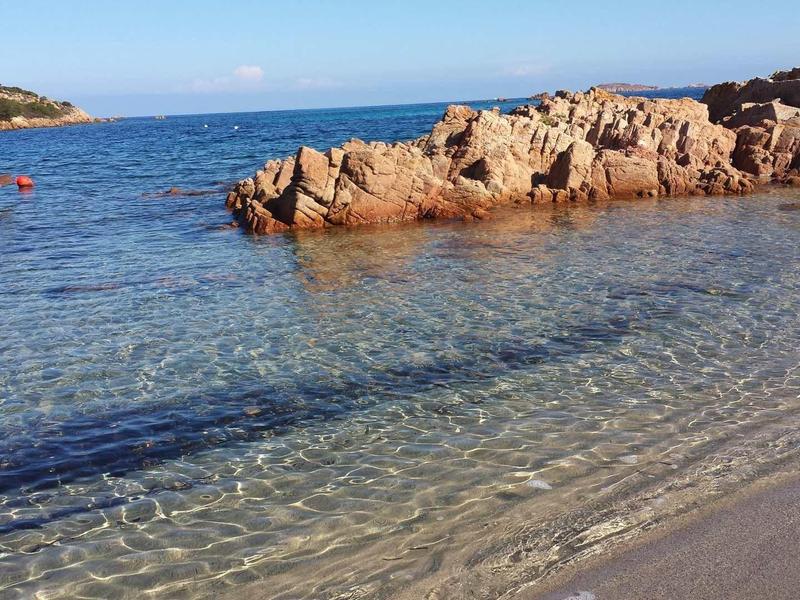 Klarer Strand mit glattem Sand, leichten Wellen und felsiger Küste im Hintergrund unter blauem Himmel.
