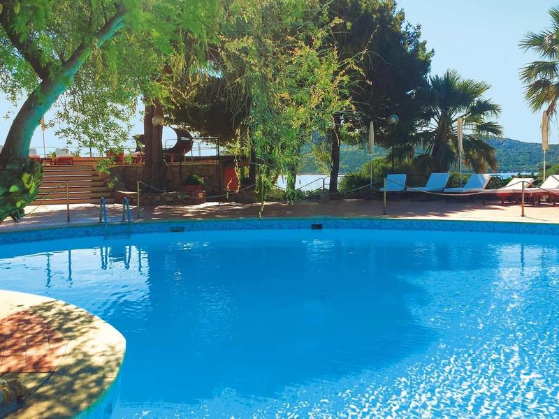 Blauer Swimmingpool mit umgebenden Bäumen und Liegestühlen unter klarem Himmel.