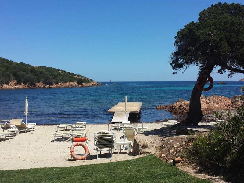 Strand mit weißem Sand, zwei Liegestühlen, blauem Meer und Baum rechts bei klarem Himmel.
